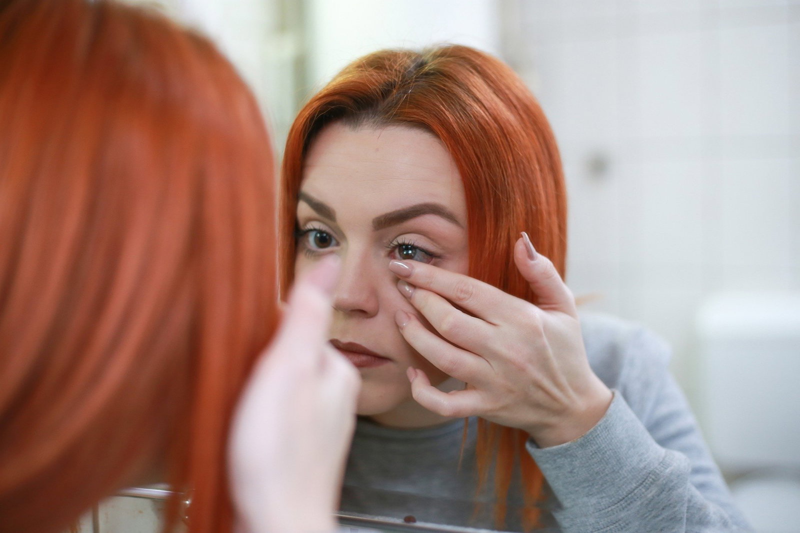 Das Bild zeigt eine rothaarige Frau im Spiegel, die sich Kontaktlinsen einsetzt.